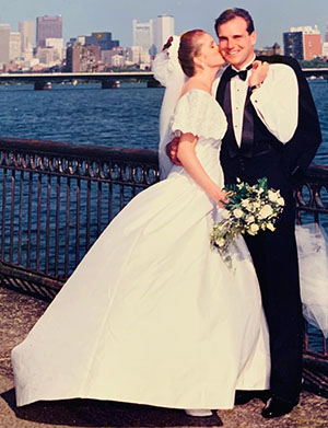 Mary Lou in a wedding dress kissing her groom, TJ, who is wearing a tux. They're standing along the waterfront in Boston.